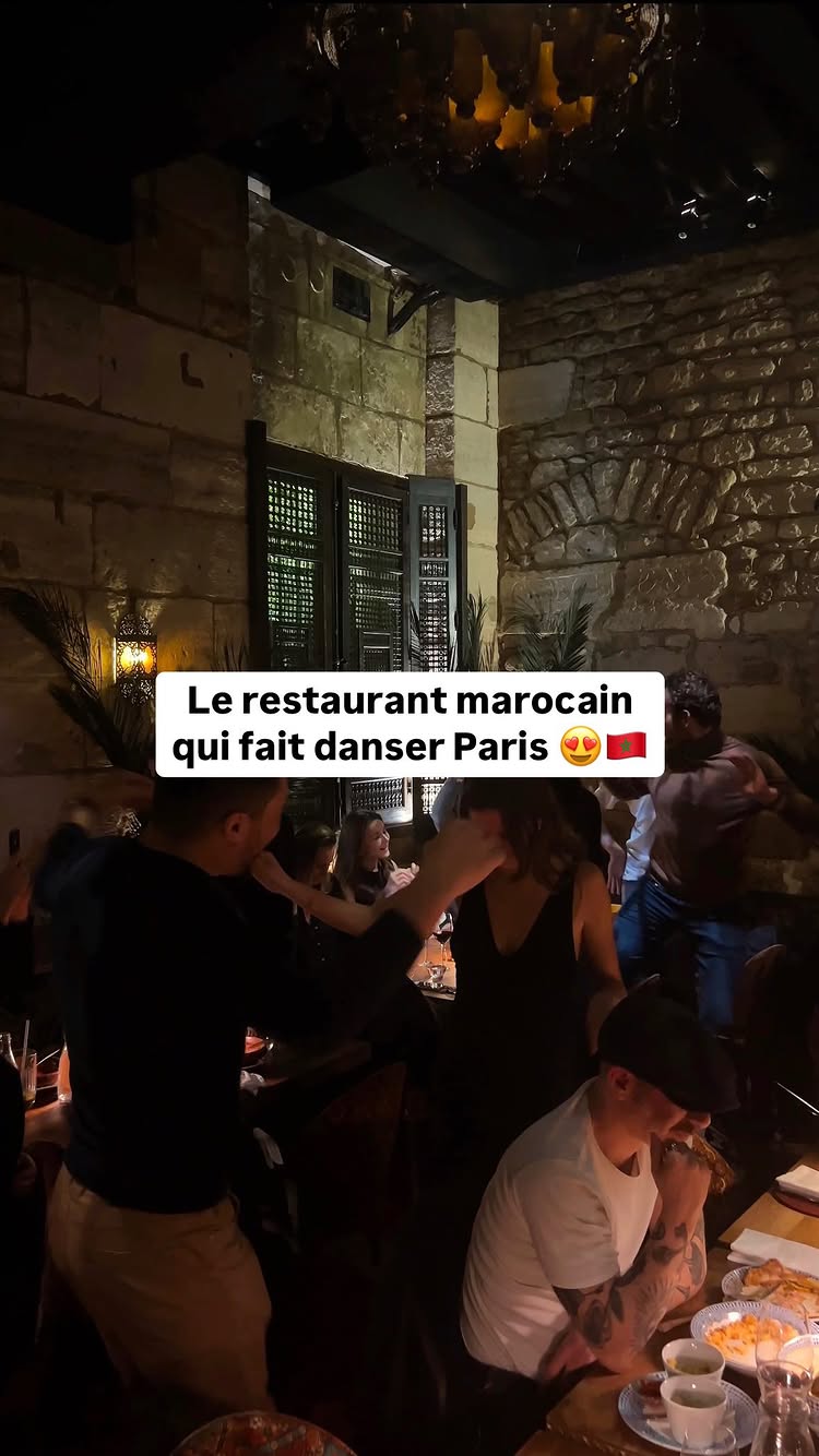 An interior shot of a dimly lit Moroccan restaurant shows groups of people gathered and dancing. In the foreground, a man in a black shirt dances with a woman in a black dress. Another man in a white shirt with tattoos sits at a table, watching. Behind them, other patrons dance near a stone wall. The lighting is warm, and the overall ambiance is festive.