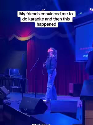 A woman stands on a stage, singing into a microphone. The background includes a red curtain and a blue spotlight from a projector. Text overlays read "My friends convinced me to do karaoke and then this happened" and "Wait for it." There are speakers and stage equipment visible.