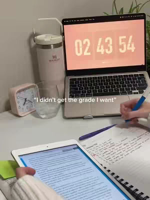 A desk setup with a laptop displaying a clock, a water bottle, a pink alarm clock, a glass, and a notepad where someone is writing with a pen. An iPad is also in the frame.