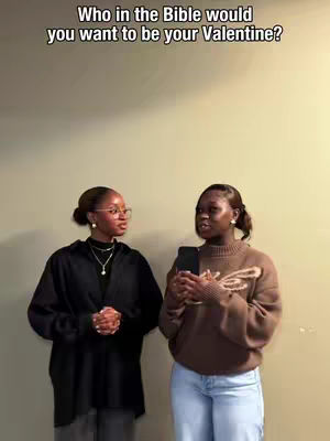 Two young women stand and face each other, appearing engaged in conversation. The woman on the left side wears glasses, a black shirt, and holds her hands clasped. The other woman holds a phone and wears a brown sweater. The background is a solid, neutral color.