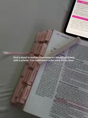 The image shows a Bible open on a flat surface, possibly a bed or tabletop. The book is open to a page with text, and has pink tabs marking certain sections. A tablet displaying text with pink highlighting is visible in the background, and a pink ribbon lies across the open pages. The lighting appears soft and diffused.