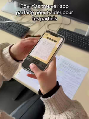 A person holds a smartphone displaying a document on a desk. The background includes a keyboard, monitor, and papers scattered around.