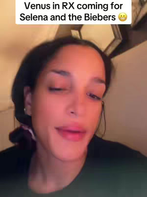 A close-up shot of a woman's face, slightly angled to the left. She has dark hair, pulled back, and is wearing a black top. Overlayed text on top reads 'Venus in RX coming for Selena and the Biebers', with a smiling emoji. The background is dimly lit, suggesting an indoor setting.