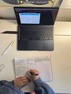 An overhead shot of a desk with a laptop, notebook, and pen. The laptop is open, displaying a document. A person's hands, with a ring on a finger, are visible writing in the notebook. A pen and highlighter are on the desk.
