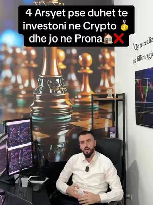 A man in a white shirt sits in an office setting, likely discussing crypto investments. Behind him is what looks like a chess board or chess pieces, and on either side of him are computer monitors displaying investing charts. The backdrop seems to be adorned with artistic gold and black tones. He is looking directly at the camera.