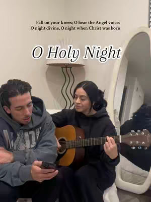 A couple is seated, with the woman playing a guitar and the man looking at his phone. Overlayed text reads "O Holy Night". The background includes a white curved structure and neutral walls, lit with soft lighting.