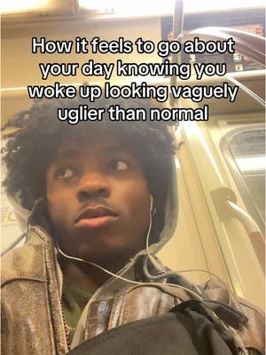 A young Black man with curly hair is seated on a train. He's wearing a brown leather jacket over a hoodie, and white earbuds are in his ears. The image has text overlayed at the top that reads, "How it feels to go about your day knowing you woke up looking vaguely uglier than normal". The man has a thoughtful expression, looking slightly to the side as if lost in thought.