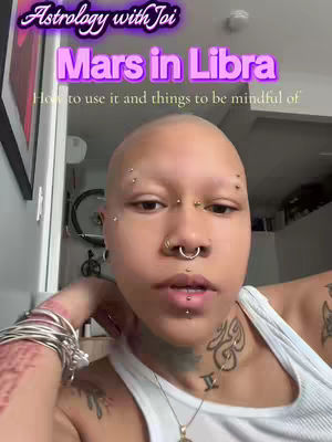 A selfie-style shot featuring a person with a shaved head, multiple piercings, and tattoos. The person is in a white tank top, with a hand resting on their face. The background is a room with visible artwork and furniture.