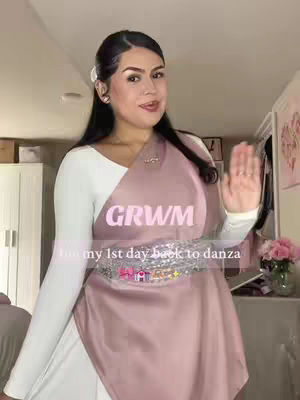 A woman with dark hair and makeup stands in a room, appearing to film herself. She wears a white long-sleeved top under a draped, mauve-colored wrap, and has a sparkly belt. Text overlays read 'GRWM' and 'for my 1st day back to danza'. The background features a white door and a few interior decorations, suggesting a personal space.