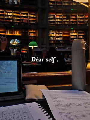 An interior shot of a library or study area, filled with bookshelves in the background. In the foreground is a desk with open notebooks and papers, a tablet, a water bottle, and a small pillow. Text overlayed on the image reads 'Dear self'. The lighting is dim, creating a warm, studious atmosphere.