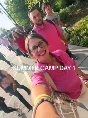 A selfie-style photo taken from a low angle, showing a group of young people and adults walking on a paved surface. The foreground features a woman with glasses smiling at the camera, with a colorful bracelet on her wrist. Behind her are several people, some wearing pink shirts and other casual attire. The environment appears to be outdoors with green foliage visible.