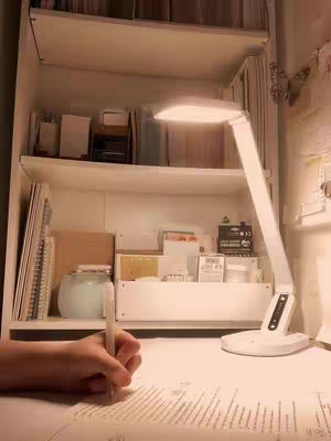 The cover image displays a well-lit desk setup focused on someone's hand writing on a sheet of paper. To the right is a white desk lamp, providing direct light. The background includes a white shelf filled with books, stationery, and other items, creating a tidy and organized study environment.