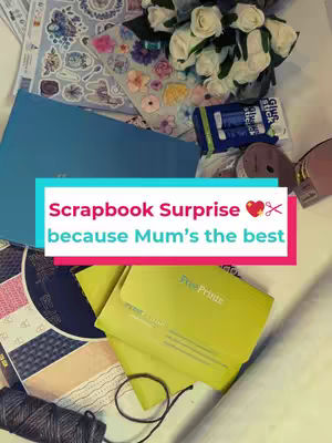 A top-down shot of various craft supplies and a partially constructed scrapbook. The items are arranged on a white surface, including a blue scrapbook, stickers, white flowers in a vase, glue sticks, and a green FreePrints booklet. The text overlay reads "Scrapbook Surprise because Mum's the best".