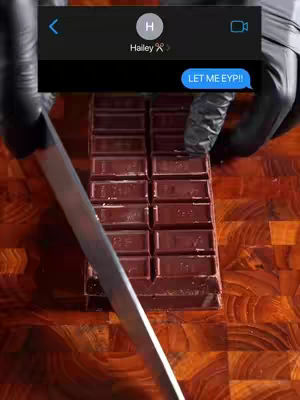 The image shows a close-up shot of a wooden cutting board with a rectangular block of chocolate being sliced by a large, silver knife. The action suggests a preparation or cutting of the chocolate slab. The image is topped with a text message overlay from a phone.