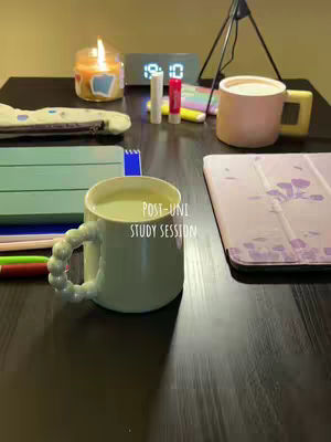 A well-lit, aesthetically pleasing desktop setup featuring a green mug with a unique handle, a lit candle in a glass jar, an illuminated digital clock, a tablet with a pink case, and various study supplies scattered on a dark wooden surface. The composition suggests a study environment with a cosy and organized ambiance.