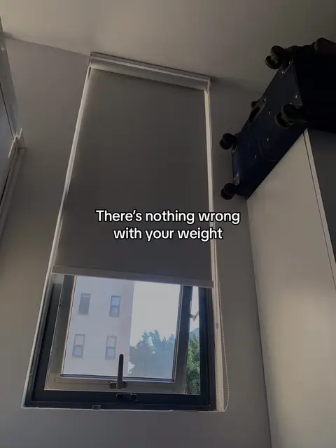 An upward-angled shot shows a window with a closed blind, a window open with the view of other buildings, and a suitcase set on top of a cabinet. The text overlay reads 'There's nothing wrong with your weight.'