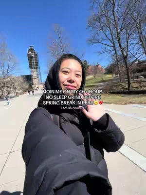 A selfie-style shot with a young woman as the primary subject. She is smiling towards the camera while holding the camera in selfie mode and walking along what appears to be a pathway with trees and buildings in the background. The sky is bright blue. She is wearing a black puffy jacket and has dark hair partially covered by a hood.