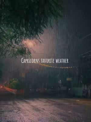 A moody, dark image captures a rainy street scene at night. Rain streaks across the image, and a street and buildings are in the background. The text overlay reads, 'CAPRICORNS FAVORITE WEATHER.' The composition is muted, with a focus on cool tones and the atmospheric effect of the rain.