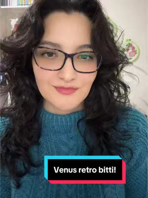 A woman with dark curly hair and glasses is centered in the frame. She wears a blue sweater, and a graphic with text overlays the bottom of the shot.