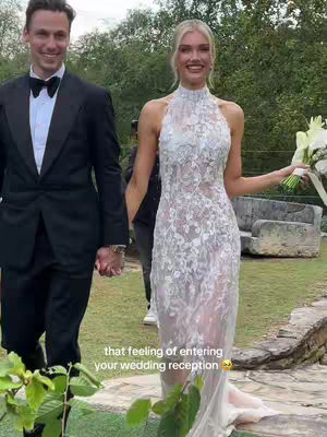A couple holds hands and walks forward on a lawn. The woman is wearing a lace wedding dress and carrying a bouquet of white flowers. The man wears a black tuxedo. Text overlay at the bottom says, 'that feeling of entering your wedding reception'.