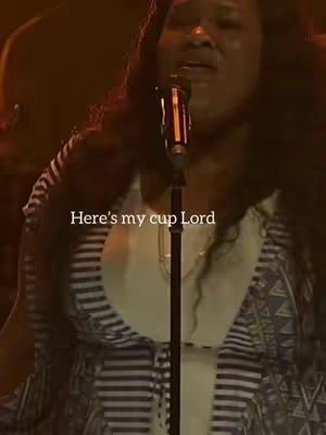 A woman with dark curly hair is singing into a microphone. She is wearing a white top with a geometric patterned vest over it. The background is dark and indistinct, suggesting a stage or performance setting.