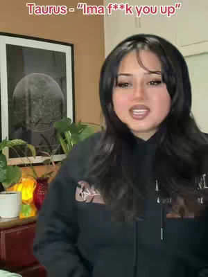 A woman in a black hoodie is shown speaking to the camera with a neutral expression. Above her head is text in a red bar. In the background, there is a framed picture of a moon with a houseplant in front of it.