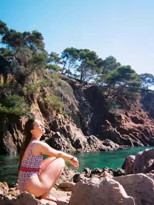 A woman in a patterned swimsuit sits on rocks near the turquoise ocean. The sunlight seems to be coming from the right. A rocky cliff with lush green trees makes up the background.