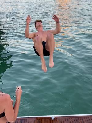 A man is mid-air, seemingly diving. The background is water, with the sky and some buildings in the distance. The man is shirtless, wearing black shorts and has both arms up. There's a wooden board in the bottom of the image, with a woman's arm visible.