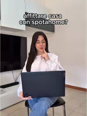 A young woman sits on a chair holding a closed laptop, with a finger to her lips as if pondering. The background includes a modern interior with a television and minimalist decor.