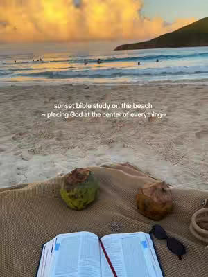 A beach scene with a sunset in the background. In the foreground, an open bible, two coconuts, and sunglasses are placed on a towel. The sky is filled with orange and yellow hues.