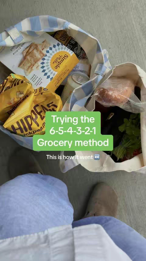 The image shows a person's legs and feet, with a grocery bag in the foreground filled with various food items. The bag is off-white with blue stripes. The items in the bag include snack foods, crackers, and other groceries. A green rectangle with white text overlays the bottom of the bag, which reads, 'Trying the 6-5-4-3-2-1 Grocery method,' and at the bottom it says 'This is how it went' with an arrow icon. Some feet and leg area in light blue shorts are clearly visible.