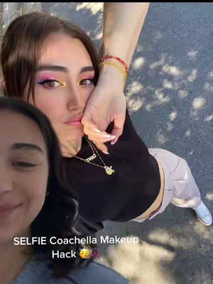 The cover image is a selfie taken at an angle, featuring a young woman in the foreground, with another person partially visible in the bottom left. The main subject has striking, colorful makeup, with pink and yellow eyeshadow, posing holding her hand near her face. She wears a black top and a necklace, set against a background of concrete with some greenery casting a shadow. Text at the bottom overlaid says 'SELFIE Coachella Makeup Hack'.