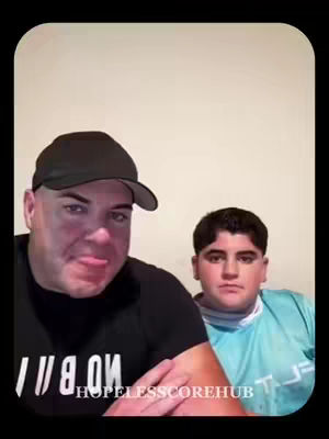 Two males are positioned in front of a white wall. One, an adult with a black cap, is making a face. The other, a younger person, is looking straight at the camera. The bottom of the image mentions a URL 'HOPELESSCOREHUB'.