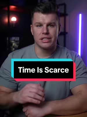 A man is centered in the shot, looking directly at the camera, with a text overlay that reads 'Time Is Scarce.' He is wearing a gray t-shirt and sitting in front of a background with blurred purple neon lights and dark furniture. His hands are clasped in front of him. The overall composition is well-lit and directs focus to the subject, making the text visually prominent.