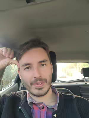 A man is sitting in the driver's seat of a car, looking at the camera. He has short brown hair, a mustache and a light beard. He is wearing a black jacket over a plaid shirt and a button-up shirt combo. The interior of the car, particularly the headrest, is visible.