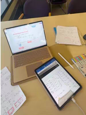 A top-down shot of a desk shows a laptop and a tablet open with what appears to be engineering school notes with various colorful writing utensils and papers scattered around.