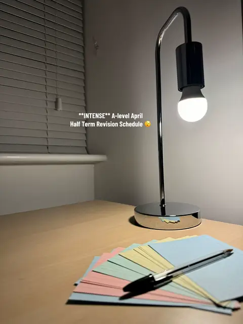 A photograph of a desk setup, illuminated by a modern desk lamp with a silver base and a black head. A stack of colorful notecards and a pen are neatly arranged on the desk's surface. The background features a window with blinds.