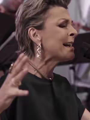 Close-up shot of a woman with short, styled gray hair, angled to the right. She's wearing a black top and large silver earrings. Her mouth is open as if singing, and her right hand is raised in a gesture. The background is slightly blurred, suggesting a live performance or recording setting.