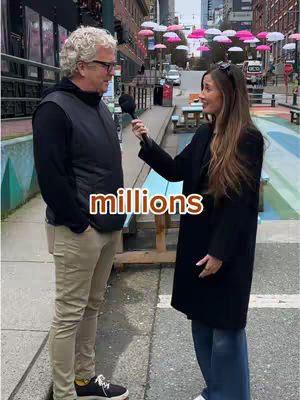 A street interview scene features a woman interviewing a man. The man, standing on the left, is wearing a black vest over a black shirt, beige pants, and sneakers. The woman, holding a microphone, wears a black coat over her shoulders with blue jeans. The background shows a city street with a series of shops and colorful umbrellas in a bright sky.