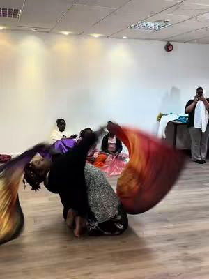 A woman is kneeling on a wooden floor, dancing with flowing colorful fabrics. Several other individuals are in the background, some sitting and others standing. The space appears to be a dance studio or similar room with minimalist white walls, and a large overhead light.