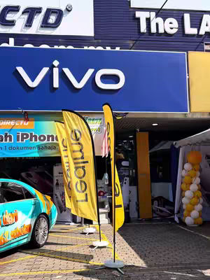 The image captures an outdoor scene with a store facade in the background. The store, displaying 'vivo' in large white letters on a blue background, suggests a mobile phone or electronics retail outlet. Two yellow feather flags are positioned in front, bearing the logo of 'Leavelive'. To the left, a portion of a car with a colorful wrap is visible. The overall composition is bright, with good lighting.