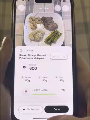 A smartphone displays a food item and its nutritional information. The phone is held by a person whose hand is partially visible. The screen shows a plate of food with meat, shrimp, mashed potatoes, and asparagus, analyzed by an app. The app shows the number of calories, protein, carbs and fat. The overall layout is well-lit, with a background of a textured surface suggesting a household setting. The app interface is clean, white, and provides data in a clear and structured manner. The phone's camera display clearly illustrates the item.