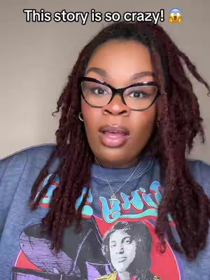 The image is a close-up of a woman with dark, curly hair, wearing black-framed glasses and a blue sweatshirt. The sweatshirt features a graphic of a person on a stage. The woman has an open mouth as though she is speaking or reacting to something. The background is a plain, light color.
