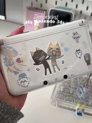 The cover image shows a close-up of a hand holding a white Nintendo 3DS console. The console is decorated with various stickers of cartoon characters, including two cat-like figures, a jellyfish, and other cute illustrations. The background is blurred, but hints at a desk setting. The overall composition is clean and the color palette is soft, with pink and white tones.