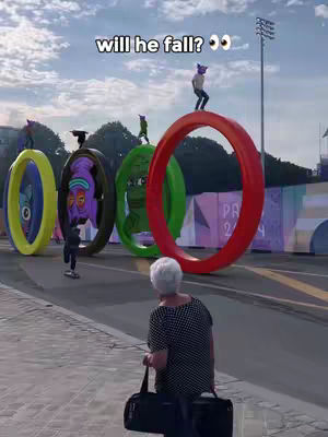 An elderly person watches in a blurry foreground as several people balance atop large, colorful ring structures, with surreal cartoon faces painted on their inner surfaces. Text appears above the scene and the setting suggests a public park or event.