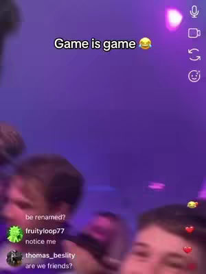 The image shows a blurry, low-angle shot of a group of people in what appears to be a dimly lit club or event setting, with a purple hue dominating the background. Above the central figures, white text says "Game is game" with a smiley face emoji. There are social media interface elements like hearts and a microphone symbol along the right side of the frame, indicating a live streaming or sharing context. The faces of people in the lower half are partially visible.