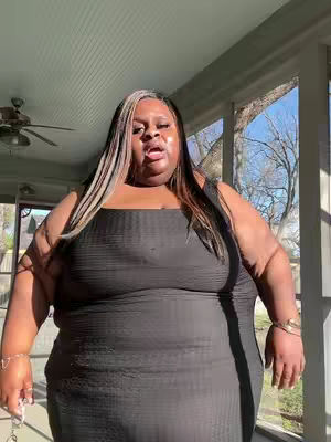 A medium shot of a Black woman standing indoors near a window. She is wearing a black dress and has long hair with blonde highlights. The woman is looking up and slightly to the left with her mouth open. The background is a blurred combination of indoor and outdoor elements.