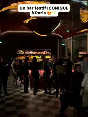 The image captures a festive, crowded bar interior, likely at night. The background features a well-stocked bar with bottles of liquor illuminated, and the foreground shows a lively crowd of people. The lighting appears to be warm, and the overall atmosphere suggests a fun, social gathering. A text overlay at the top of the image reads, 'Un bar festif ICONIQUE à Paris', identifying the location as an iconic party bar in Paris.