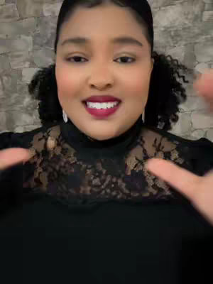A medium shot features a woman with dark curly hair styled in two small pigtails. She has a warm skin tone, wearing dark lipstick, and a black top with a lace overlay. Her fingers are spread out slightly in a gesture, and she is smiling with perfect white teeth. The background is a textured gray.