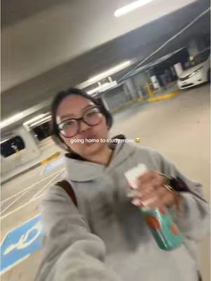 A selfie-style shot of a person walking in a parking garage or similar structure. The person is wearing glasses, a grey hoodie, and carrying a water bottle. The environment seems to be a bit sterile with concrete and fluorescent lighting.